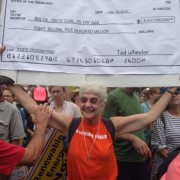 Sandy Polishuk Protesting white woman, gray hair, red t-shirt, smiling, holding up sign (illegible)